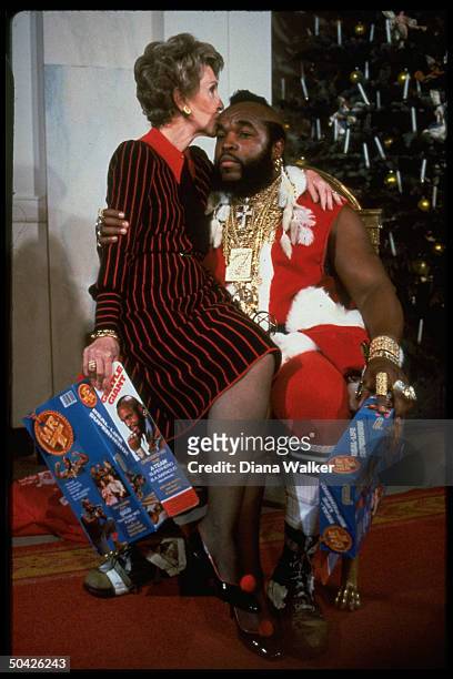 First lady Nancy Reagan sitting on lap & kissing Mr. T done up as Santa Claus at WH Christmas decoration tour.