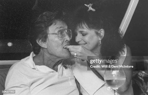 Singer, businessman Jimmy Dean being fed a cracker w. Cheese by his playful fiancee, singer Donna Meade, as they dine on board his yacht.