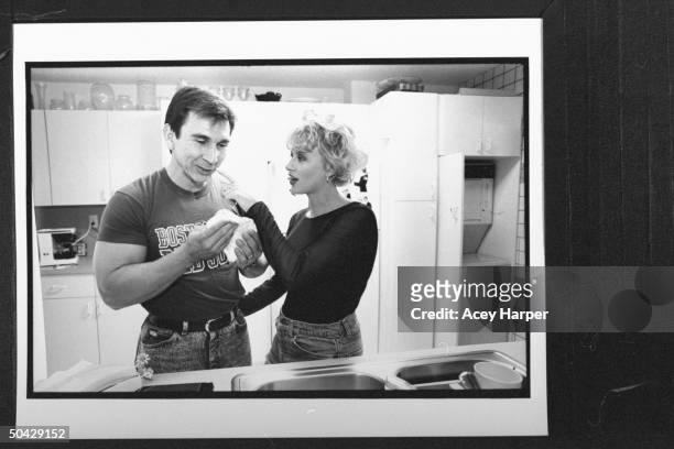Actress Victoria Jackson pointing to item on the upper lip of her husband Paul Wessel, as he holds washcloth nr. Sink in kitchen at home.