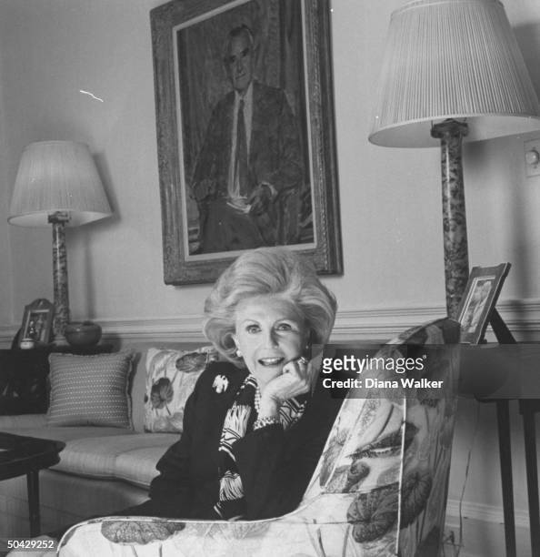 Pamela Harriman sitting in a chair at home, a portrait of her late husband, politician Averell Harriman is in the bkgrd, Georgetown.