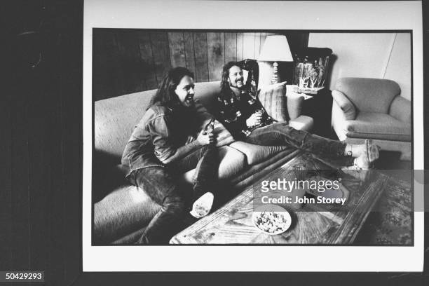 James Hetfield lead singer and rhythm guitarist for the rock group Metallica, w. Drummer Lars Ulrich, holding drinks as they relax on sofa backstage...