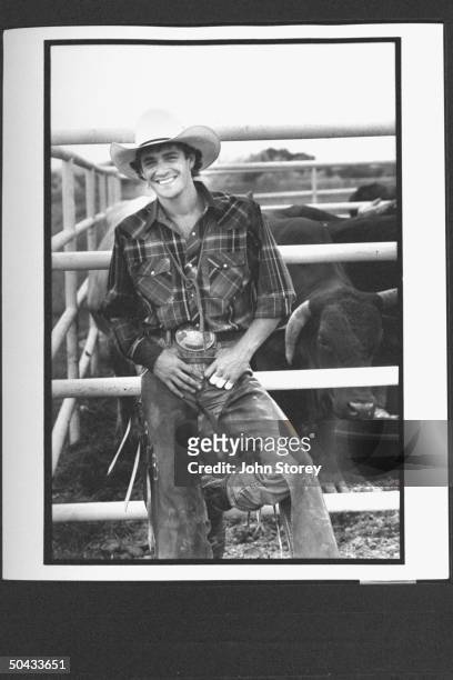Actor Luke Perry in cowboy garb, leaning against iron fence by penned bulls during a break in the filming of the rodeo movie 8 Seconds.
