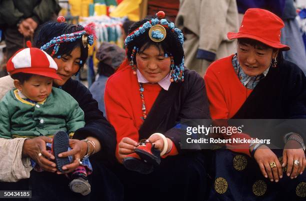Trio of Tibetan Khampa women wearing turquoise jewelry, 1 w. Child perching on her lap as another handles pair of toddler shoes.