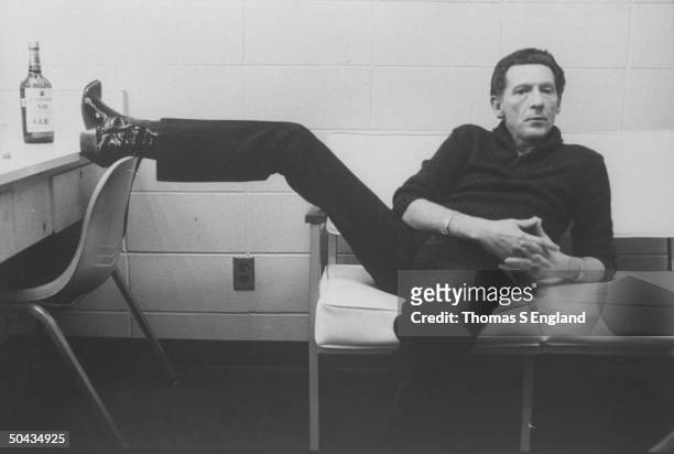 Singer Jerry Lee Lewis lounging on couch w. One foot up on dressing table next to a bottle of Seagram's V.O. Whiskey in dressing room at Performing...
