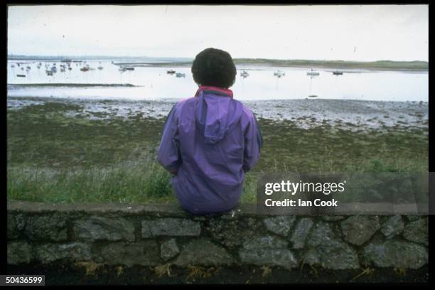 Rear view of Jenny Cockell sitting on stone wall by beach, after reunion w. The children of Mary Sutton, who she claims to have been in her past life.