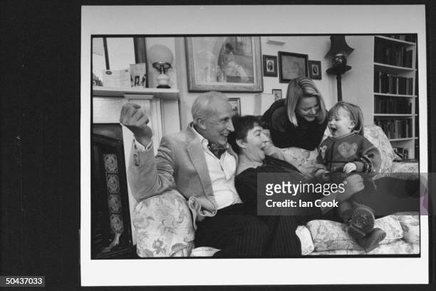 Actor Ian Richardson snuggling on couch w. Ex-actress wife Maroussa Frank who is singing w. Their granddaughter Annabel while holding her on her lap...
