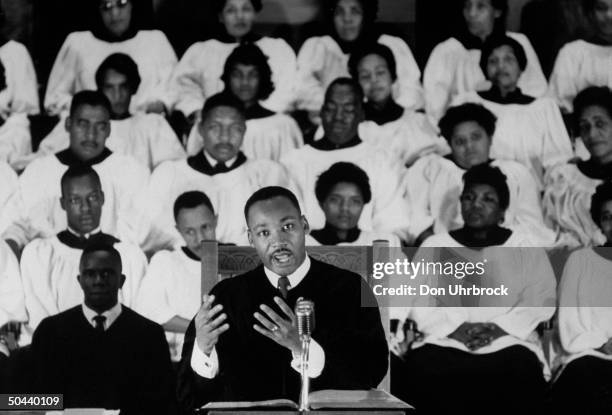 Civil Rights activist Rev. Dr. Martin Luther King Jr. Standing at pulpit delivering his sermon as a white-robed choir listens in the bkgd. At...