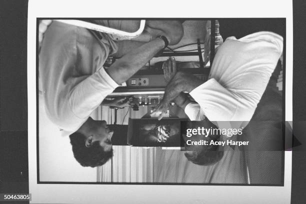 Actor Charlton Heston leaning against a film editing table and gesturing as he talks to his son, Fraser, sitting nearby, who is directing Heston in...