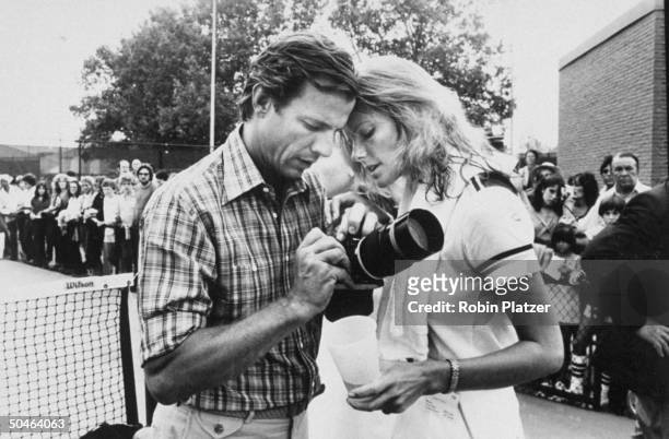 Photographer Peter Beard holding camera w. Model/wife Cheryl Tiegs on tennis court, as crowd watches behind fence.