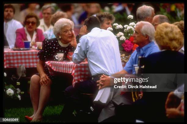 Pres. & wide-eyed Barbara Bush conferring in hushed, conspiratorial tones, at table in midst of guests, hosting WH barbeque/picnic-type fete .