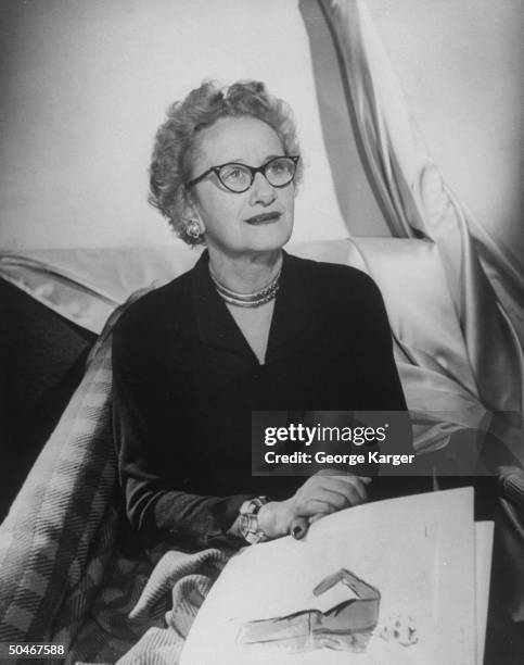 Dress designer Hattie Carnegie holding sketches in hand & sitting amid bolts of material.