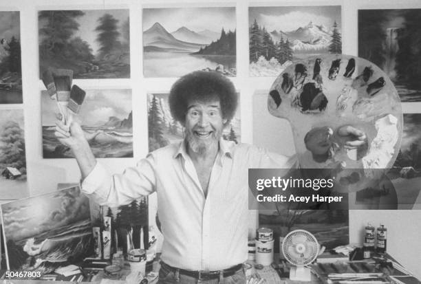Painting instructor/artist Bob Ross jubiantly holding up paint pallette & brushes as he stands in front of wall covered w. His landscape paintings in...