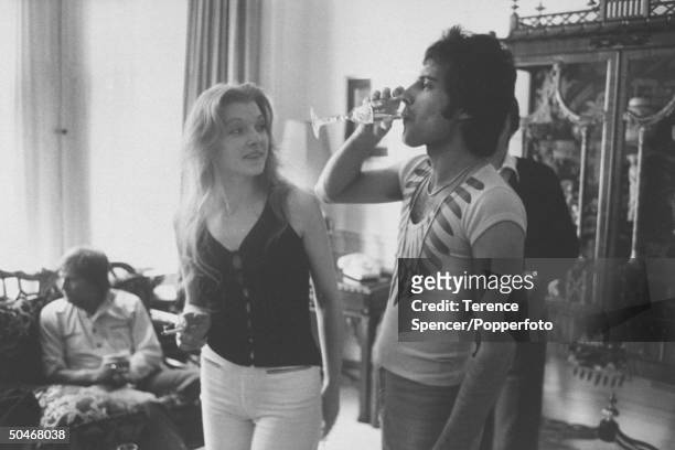 British rock singer Freddie Mercury of Queen drinking a class of champagne as his girlfriend Mary Austin looks on during party for friends at home in...