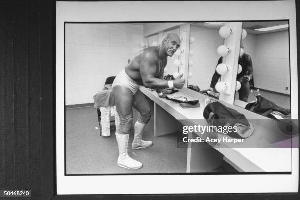 Wrestler Hulk Hogan partially dressed in his wrestling togs, giving thumbs-up sign as he leans on counter in front of mirrors in dressing room before...