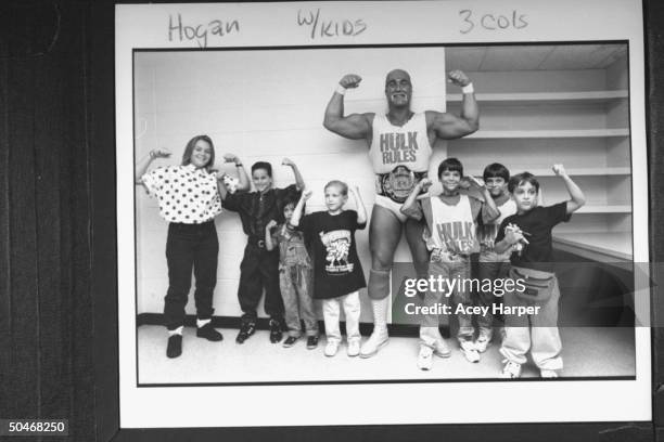 Wrestler Hulk Hogan in wrestling togs incl. His championship belt, flexing his biceps amidst 7 youngsters who are imitating his pose in dressing room...