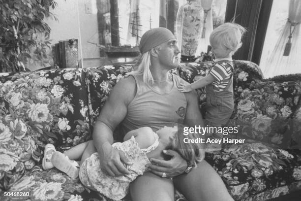 Wrestler Hulk Hogan cuddling w. His 3-yr-old daughter Brooke as he chats w. His 1-yr-old son Nicholas while relaxing together on a floral print couch...