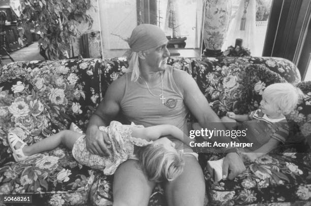Wrestler Hulk Hogan cuddling w. His 3-yr-old daughter Brooke as he chats w. His 1-yr-old son Nicholas while relaxing together on a floral print couch...