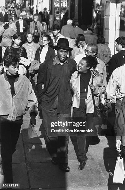 Actor Danny Glover wearing hat as he walks w. His arm around wife Asake Bomani while strolling on crowded Stockton St.