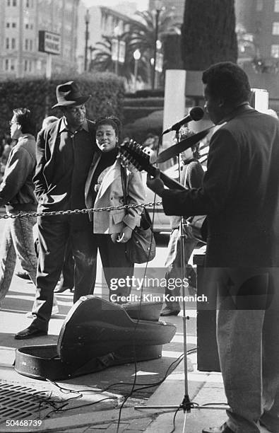 Actor Danny Glover wearing hat w. His arm around wife Asake Bomani as they stand listening to man singing into a mike & playing guitar on the street.