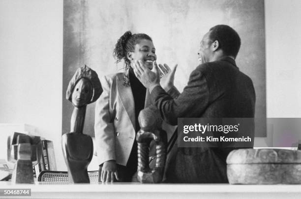 Actor Danny Glover chatting affectionately w. His wife Asake Bomani at desk w. African art objects on it at her yet-to-be completed Bomani Gallery.