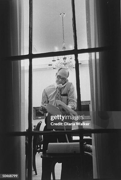 Adv. Brent Scowcroft in shirt-sleeves, talking on telephone as he goes over some papers while standing in his WH office at night as seen through...