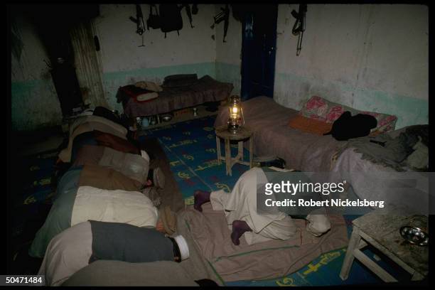 Muslims at prayers, mujahedin Salafiys, framed by wall-hung rifles, inside rm. W. Barracks-style bedding, in Salafiya rebel-held Kunar province.