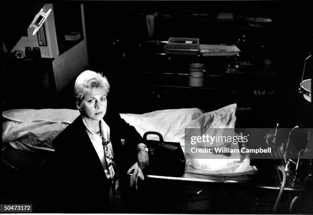 Author Patricia Cornwell holding pair of rubber gloves as she nonchalantly rests her arm on black medical bag which sits next to covered corpse w....
