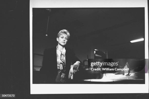 Author Patricia Cornwell holding pair of rubber gloves as she nonchalantly rests her arm on black medical bag which sits next to covered corpse w....