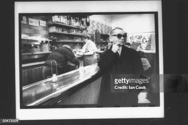 Fashion designer Karl Lagerfeld pensively fingering his chin as he stands at bar in a bistro.