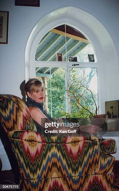 Singer Marianne Faithfull holding lit cigarette as she curls up in large chair covered in colorful bargello fabric, at her 19th C. Cottage.