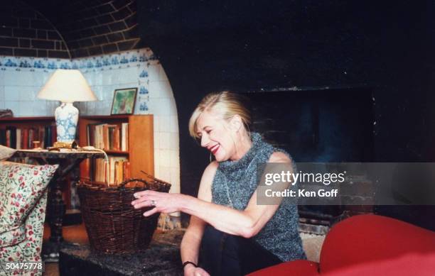 Singer Marianne Faithfull holding lit cigarette as she sits in front of peat fire burning in fireplace, in her 19th C. Cottage.