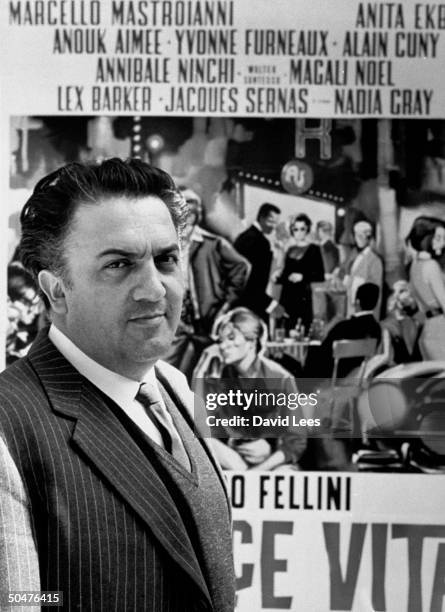 Italian dir. Federico Fellini standing in front of poster for his film La Dolce Vita.