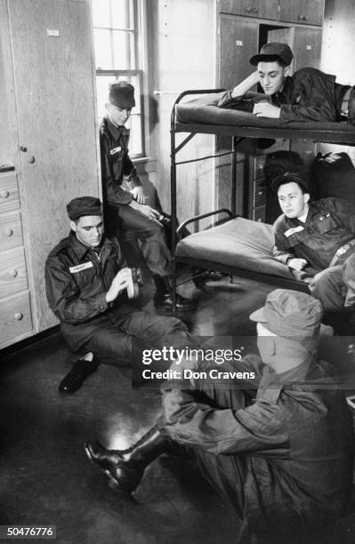 Singer/Army Pvt. Elvis Presley clad in Army fatigues & cap, sitting on barrack floor as he brush-polishes his Army shoes while 4 other inductees look...