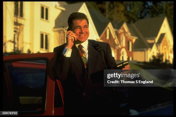 Ralph Roberts, named best-selling realtor in US, talking on cell phone while leaning on car outside in upper class neighborhood.