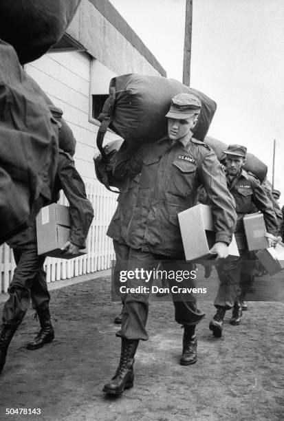 Singer/Army Pvt. Elvis Presley clad in Army fatigues & cap, w. Duffel bag over his shoulder & box under one arm, w. Several inductees as they walk to...
