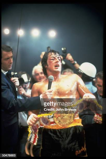 Actor Sylvester Stallone holding championship belt, surrounded by reporters, after winning fight in scene fr. Motion picture Rocky V..