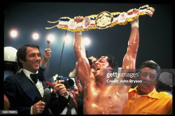 Actor Sylvester Stallone victoriously holding championship belt over head while reporters watch in scene fr. Motion picture Rocky V.