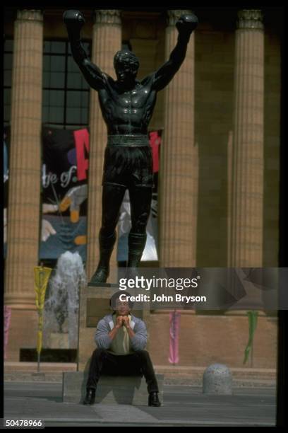 Actor Sylvester Stallone sitting under statue of Rocky Balboa, character he made famous in Rocky films.
