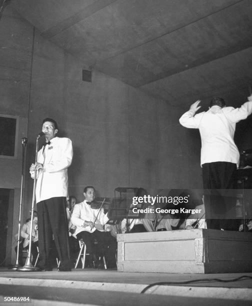 Singer Frank Sinatra rendering a song as conductor Max Steiner leads the New York Philharmonic orchestra during concert featuring the crooner which...