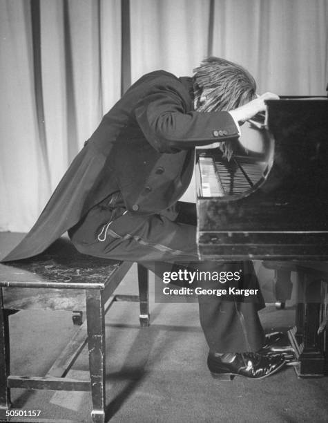 Henry Scott, wearing a tuxedo and a wig, banging his head on the wood of a piano.