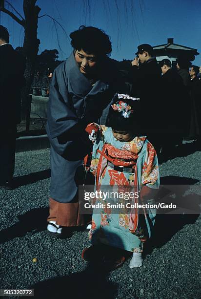 Japanese woman bending down to hold the hand of her daughter, who wears a red and blue kimono, a crowd of men in the background standing outside a...