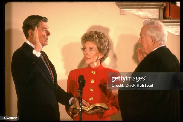 President Ronald Reagan holding up right hand and left hand on bible as wife Nancy looks on while Chief Justice Warren Burger swears Reagan in for...