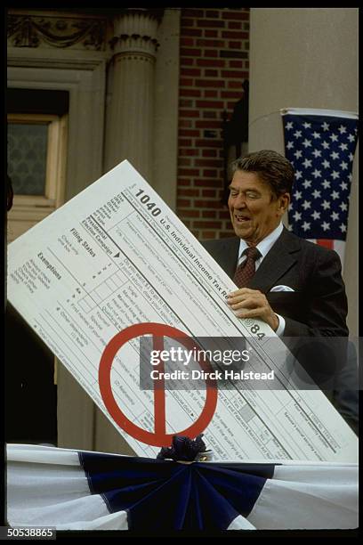 President Ronald Reagan holding up giant 1040 income tax form, emblazoned with a red circle bisected by a red line, while giving tax reform speech.