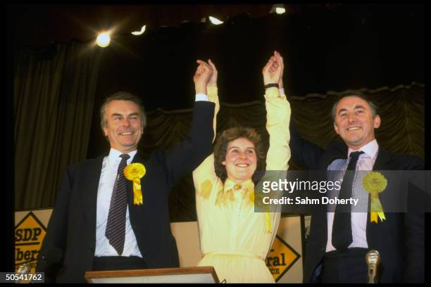 Liberal Party leaders David Steel and David Owen victoriously holding up arms of SPD/Liberal Party candidate Rosie Barnes at a rally for her election.