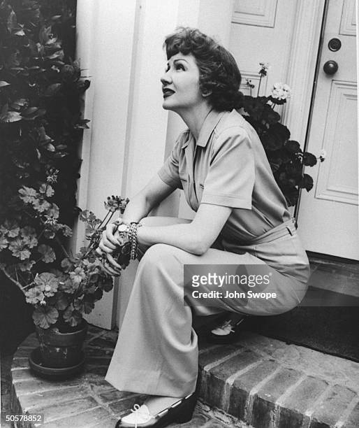 Actress Claudette Colbert wearing casual pants, sitting alone on porch steps, and thoughtfully gazing skyward.