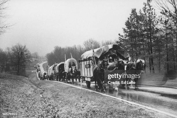 Overview of horse-drawn covered wagon train trekking up highway reenacting 1,000-mile journey that Cherokees traveled 150 years ago came to be known...