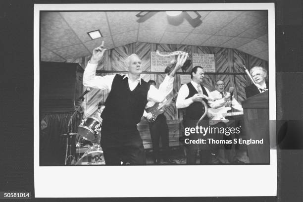 Church leader Dewey Chafin w. Ray Christian handling timber rattlesnakes while trio of musicians play & sing behind them during worship service at...