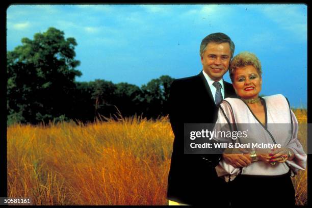 Former televangelists Jim and Tammy Faye Bakker, in expensively tailored clothes, standing in the middle of a wheat field.