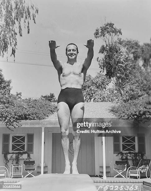 Portrait of actor John Payne wearing a pair of small swimming shorts as he prepares to use a diving board over a pool, circa 1945.