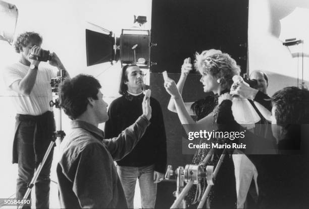 Model Cheryl Tiegs checking her hairdo as male assistant holds up new Barbie-sized, super model doll in her likeness while photographer & others fuss...